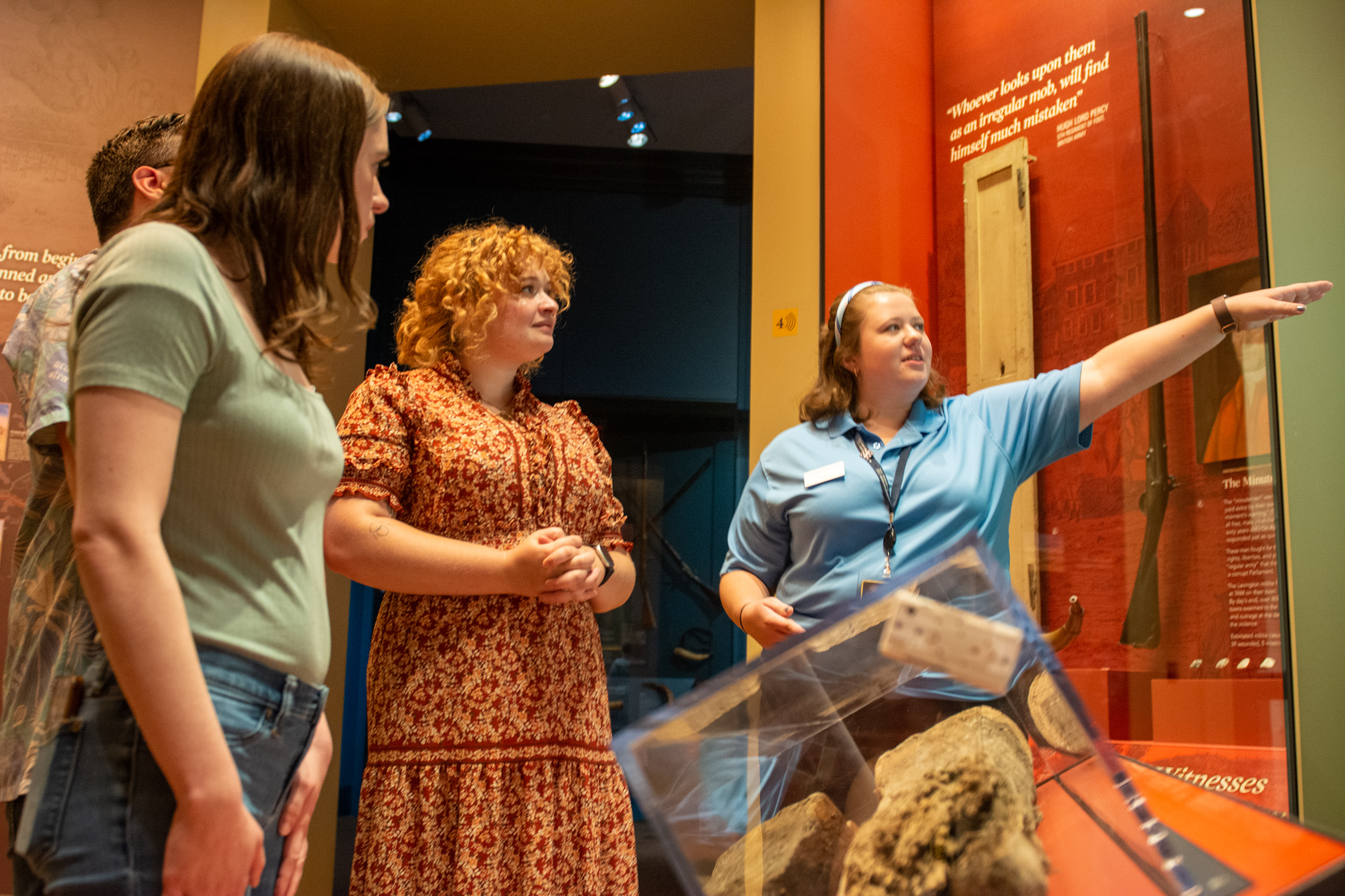 A guide in a blue polo shirt points out something in a glass case in front of a group of people