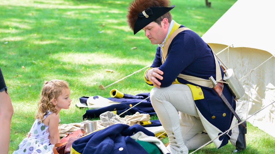 Costumed living history interpreter Joel Anderson kneels to the right of the image wearing a blue and gold commander in chief's guard jacket and blacket and fur lined hat while talking to a young visitor at Morristown National Historical Park.