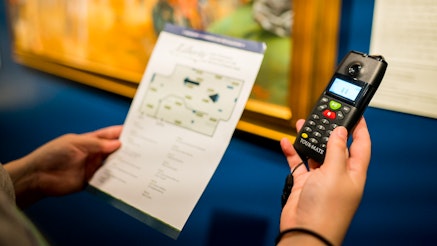 A guest holds a Museum-issued audio tour device in their right hand and the printed audio tour guide in their left.