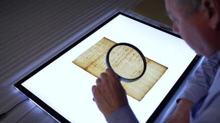 A Museum curator views a document on a lightboard with while looking through a large magnifying glass.