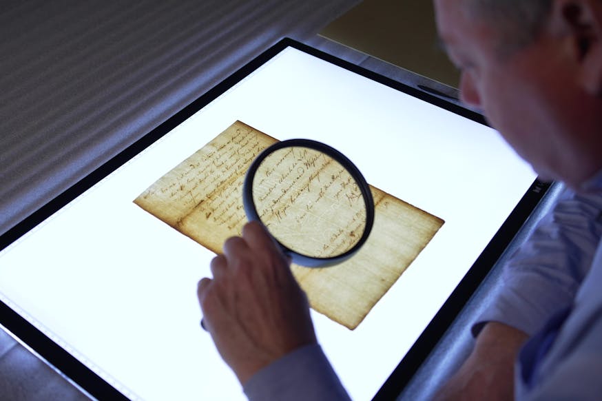 A Museum curator views a document on a lightboard with while looking through a large magnifying glass.