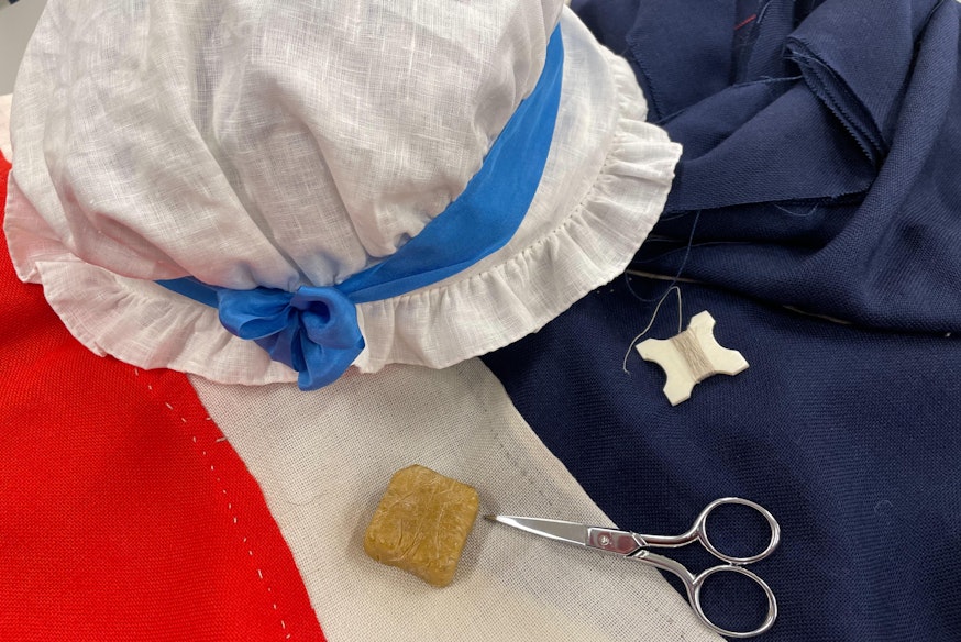 image of red, which, and blue fabric, a white bonnet, scissors, thread, and other flag making tools.