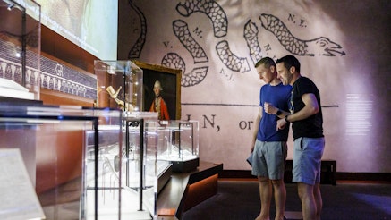 Two adult male guests wearing shorts and t-shirts look at powder horns in the Museum's Join or Die gallery.
