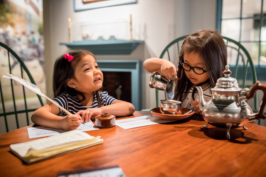 Two Asian American female children utilize the objects within Revolution Place. They are both seated at a wooden table. The child on the left is writing with a quill pen and smiling, while the child on the right is pretended to pour cream into her silver teacup. On the table to her left is a silver tea pot.