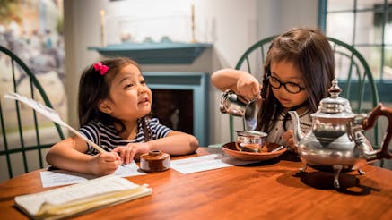 Two Asian American female children utilize the objects within Revolution Place. They are both seated at a wooden table. The child on the left is writing with a quill pen and smiling, while the child on the right is pretended to pour cream into her silver teacup. On the table to her left is a silver tea pot.