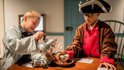 Two white male adolescents, both standing at a wooden table, interact with a play tea set. The adolescent on the left is pretending to pour tea into a silver teacup from a silver teapot. The adolescent on the right has his hand on the teacup saucer. Both are dressed in Revolutionary-era outfits.