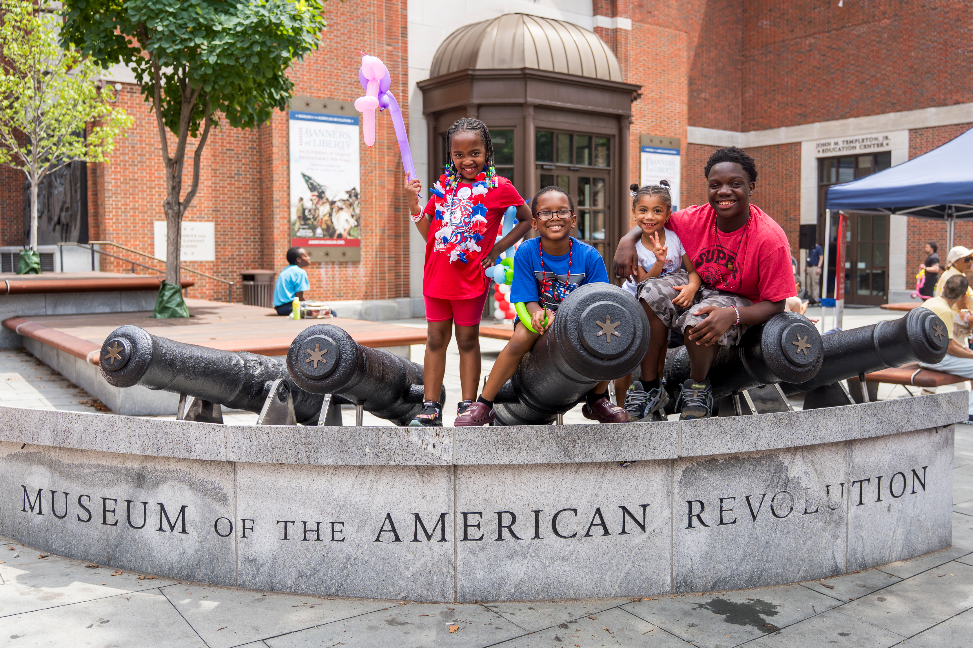 Four children of varying ages pose on top of steel cannons in front of the Museum of the American Revolution.