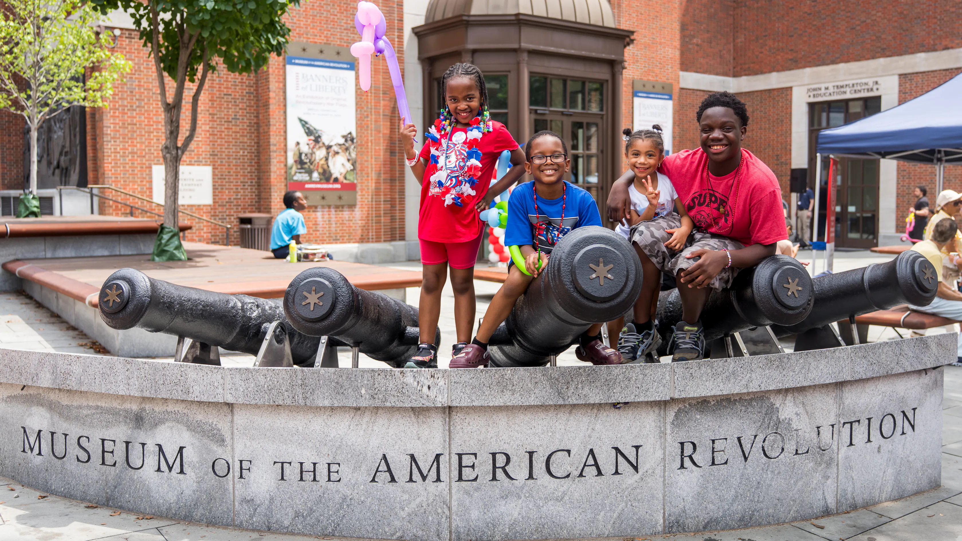 Four children of varying ages pose on top of steel cannons in front of the Museum of the American Revolution.