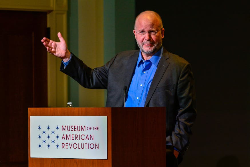 William Hogeland, a white man with glasses and a salt and pepper beard, speaks from a podium with the Museum logo on it.