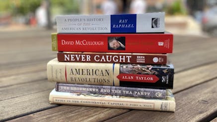 200th Edition Museum Staff Picks Reading List includes A People’s History of the American Revolution by Ray Raphael; John Adams by David McCullough; Never Caught by Erica Armstrong Dunbar; American Revolutions: A Continental History 1750-1804 by Alan Taylor; The Shoemaker and the Tea Party by Alfred F. Young; and Spies in the Continental Capital by John A. Nagy. The books are stacked on top of one another on a wooden bench outside on a sunny day. The books and the bench are in clear view while the background is blurred.