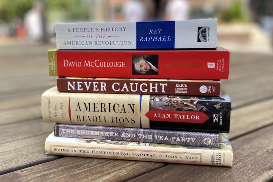200th Edition Museum Staff Picks Reading List includes A People’s History of the American Revolution by Ray Raphael; John Adams by David McCullough; Never Caught by Erica Armstrong Dunbar; American Revolutions: A Continental History 1750-1804 by Alan Taylor; The Shoemaker and the Tea Party by Alfred F. Young; and Spies in the Continental Capital by John A. Nagy. The books are stacked on top of one another on a wooden bench outside on a sunny day. The books and the bench are in clear view while the background is blurred.
