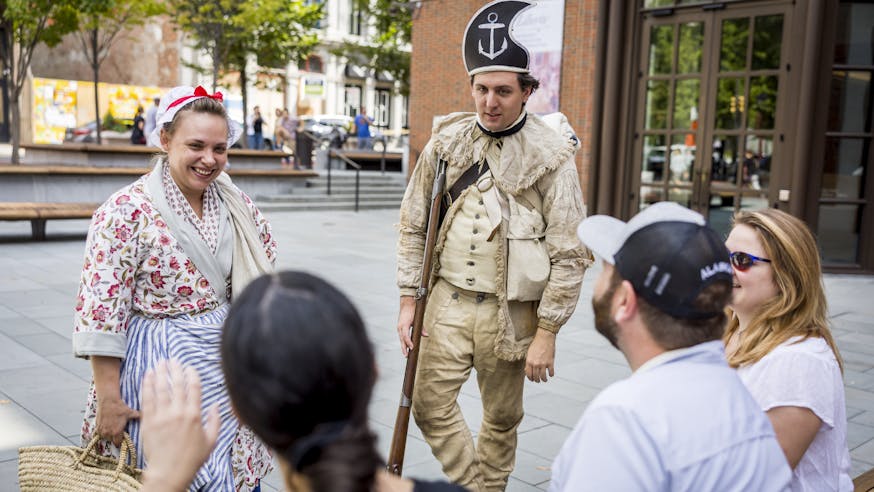 Two living history interpreters talk with a group of visitors on the Museum's outdoor plaza.