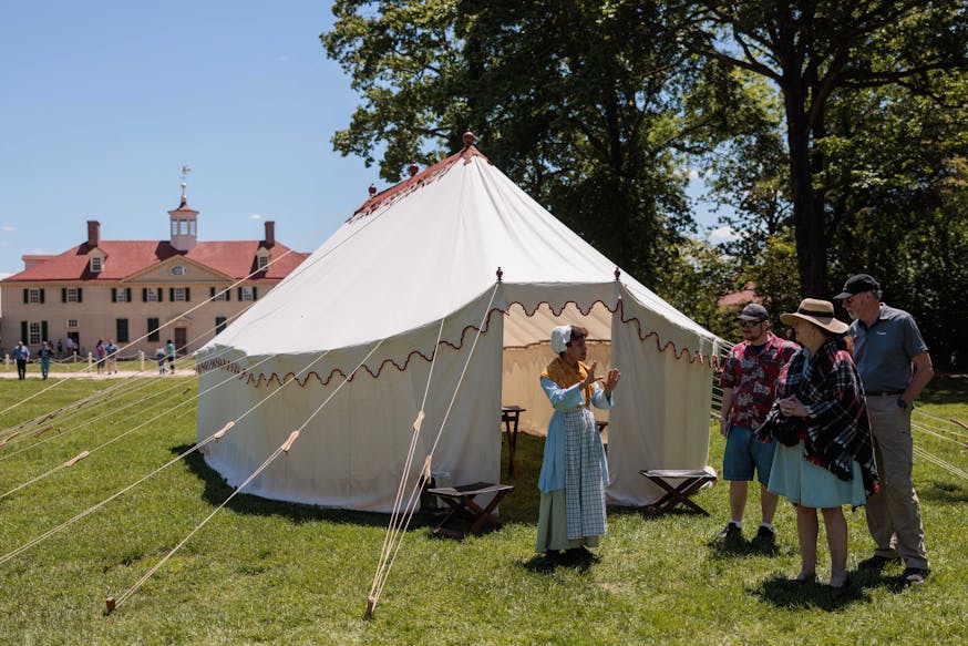 A costumed historical interpreter speaks with three guests in front of the Museum's replica of George Washington's tent on the lawn at Mount Vernon with the mansion in the background.