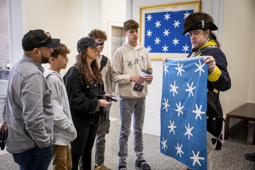 A Museum educator dressed in 18th century clothing shows visitors the Museum's handsewn silk replica of Washington's Standard flag.
