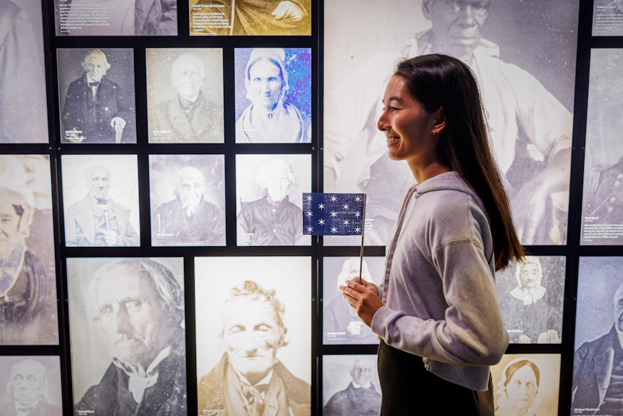 A visitor holds a miniature version of George Washington's standard flag in front of photographs of people from the Revolutionary generation who lived into the age of photography.