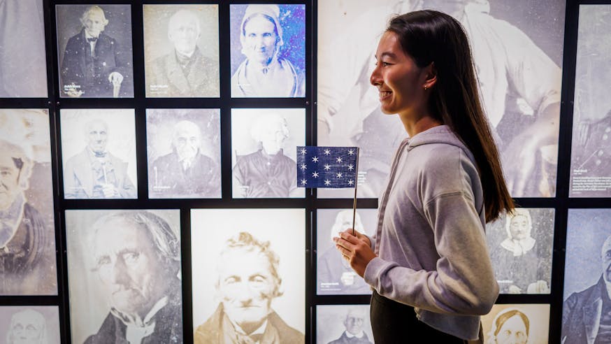 A visitor holds a miniature version of George Washington's standard flag in front of photographs of people from the Revolutionary generation who lived into the age of photography.