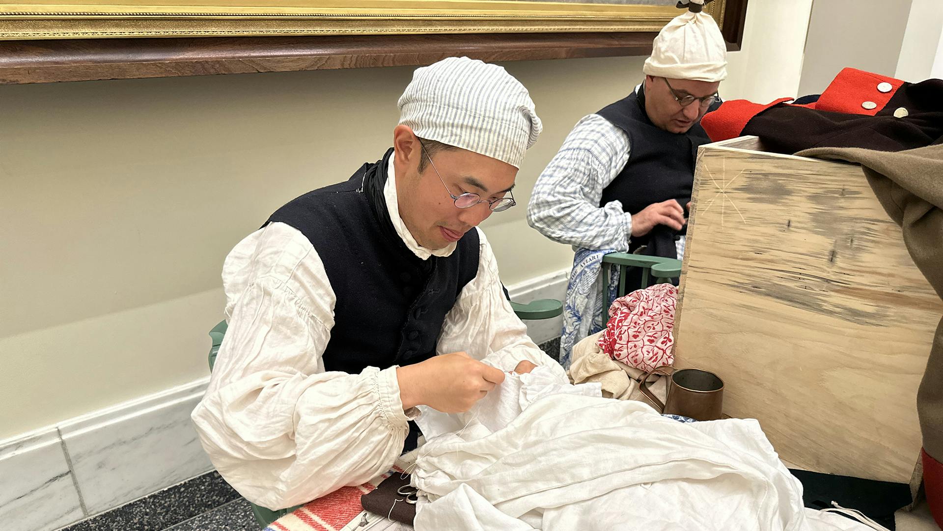 A costumed male historical interpreter in a white shirt, blue vest, and white hat sewing a white sail draped on a table.