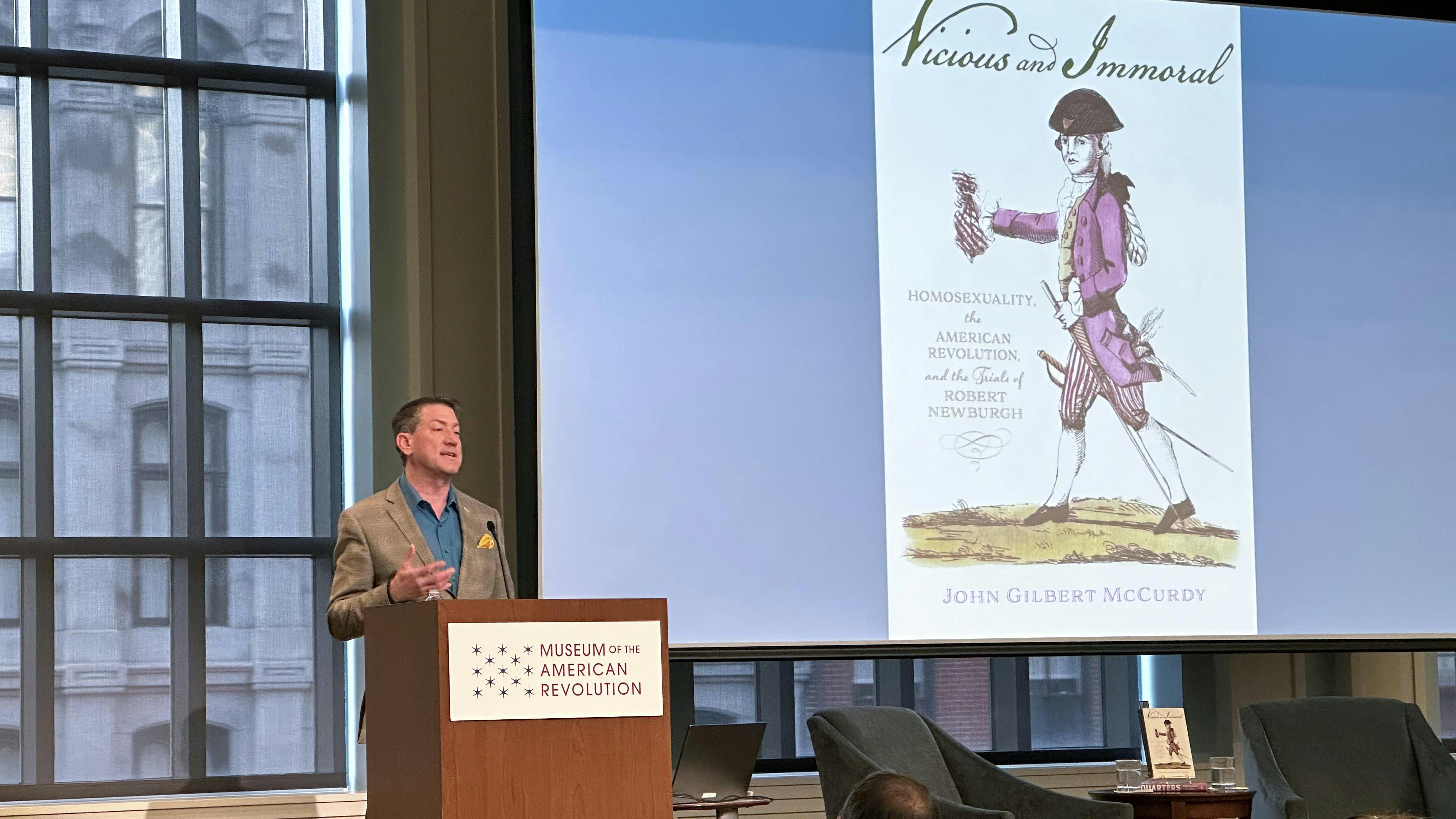 Dr. John Gilbert McCurdy stands at a podium with the Museum of the American Revolution logo and an image of his Vicious and Immoral book on the screen behind him.