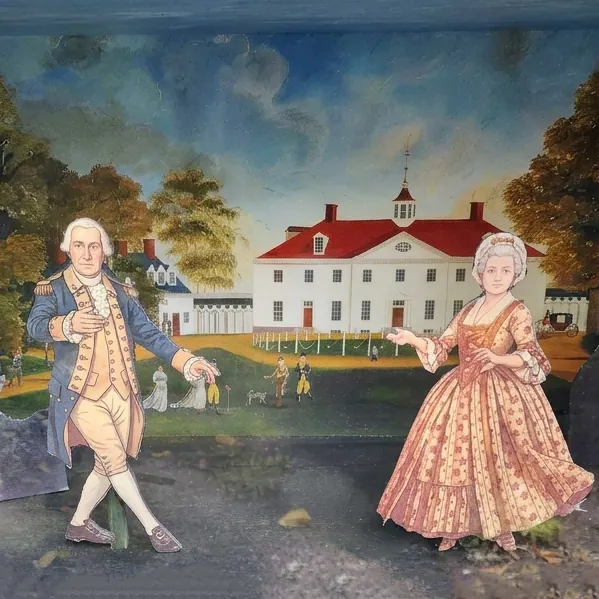 Two wooden cutouts of a man and woman in 18th century clothing in front of a painting of a landscape with a large white building