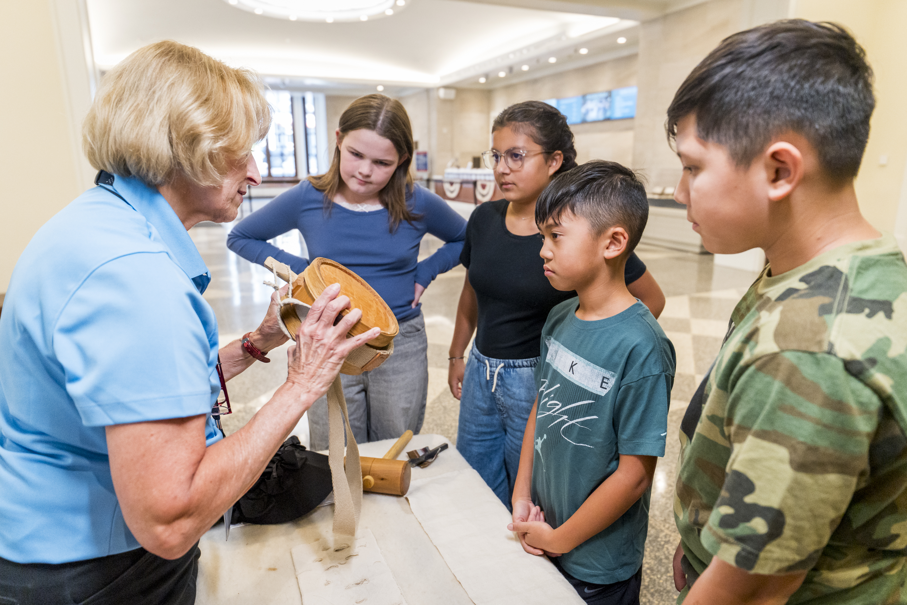 A woman in a light blue shirt shows a wooden canteen to a group of children