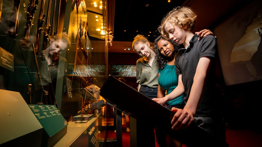 Three guests view the Arms of Independence case and touch screen in the Museum galleries.