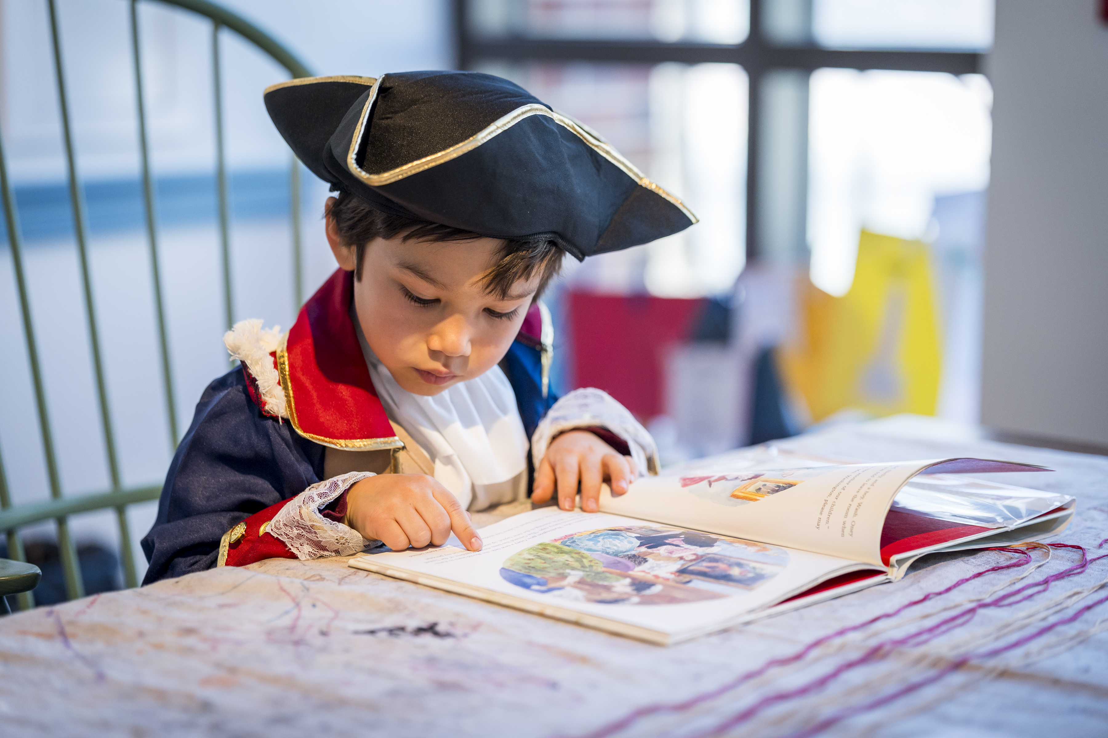 Child wearing a tricorn hat and red and navy blue coat reads a children's book