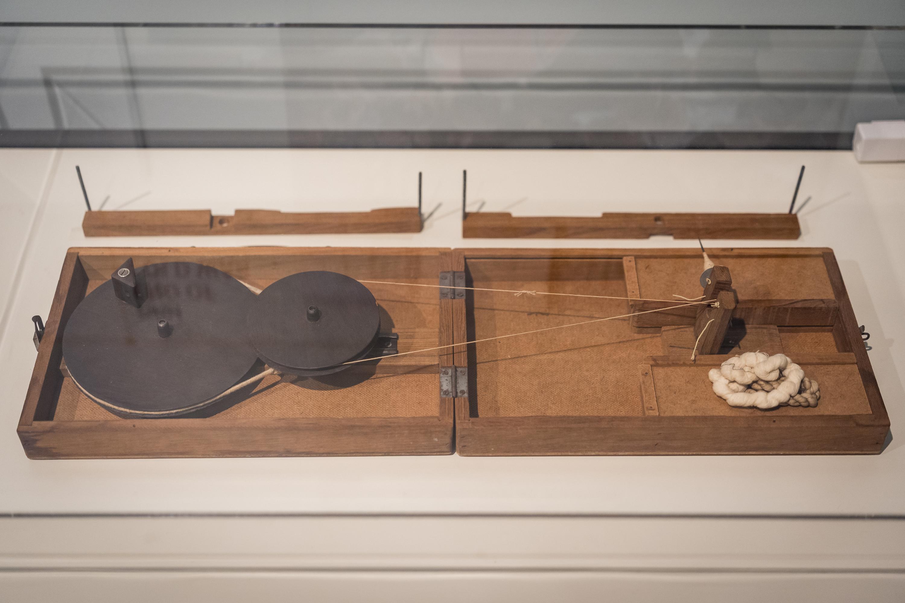 A flat wooden spinning wheel in a glass case