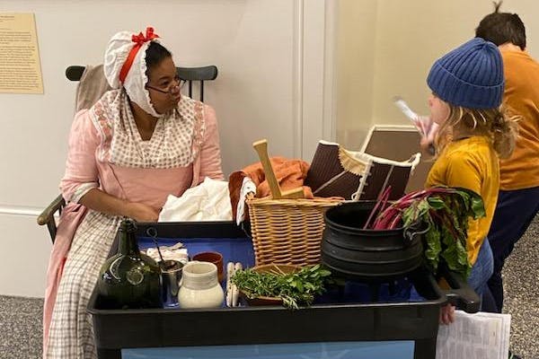 Costumed living historian Leslie Bramlett sitting in a chair at left of the image wearing 18th century pink and white dress talking to a young visitor at a museum discovery cart with replica objects.