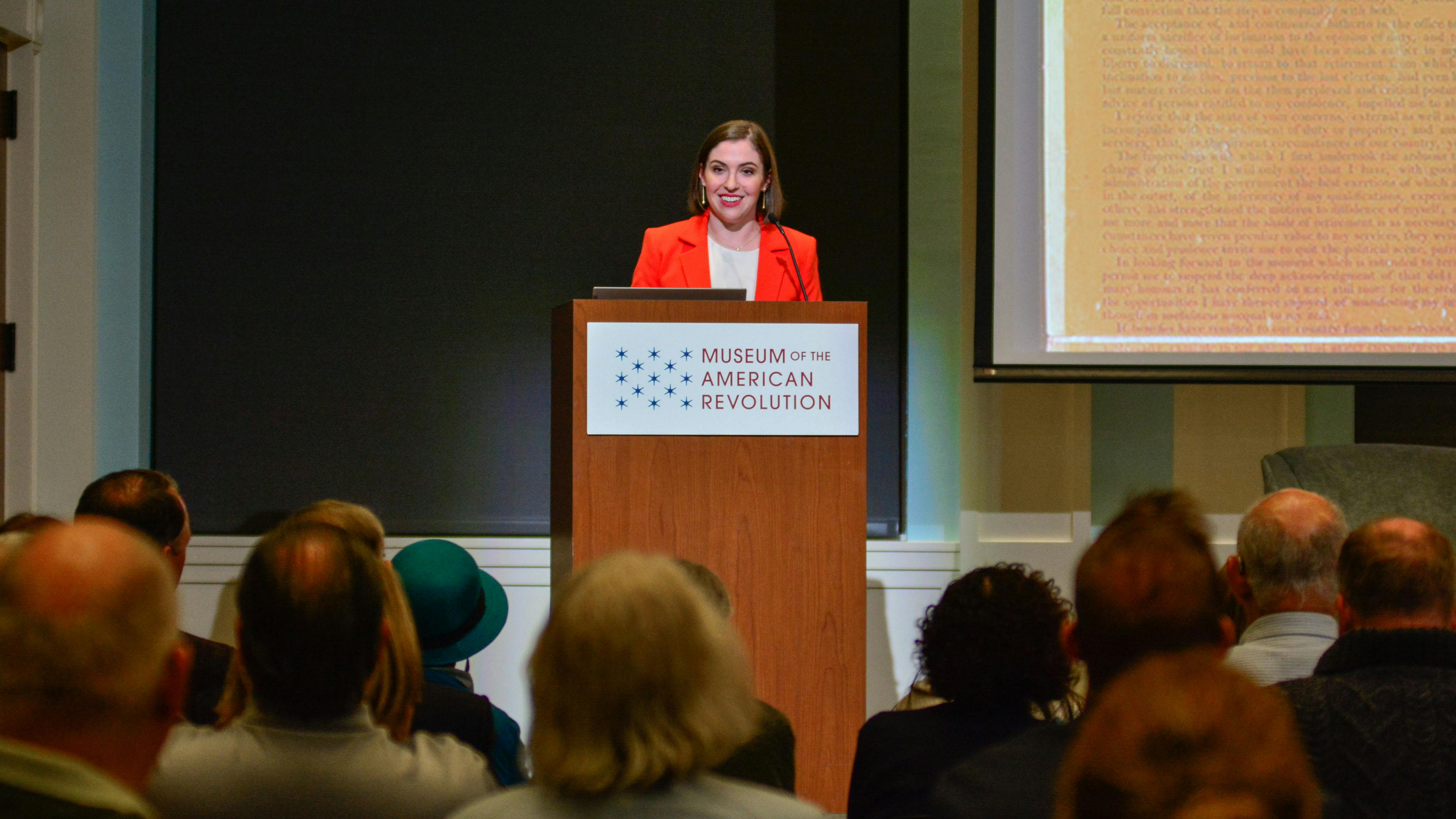 This image shows Dr. Lindsay M. Chervinsky standing at a wooden podium with the Museum's logo on the front while wearing a red suit with a crowd watching her.