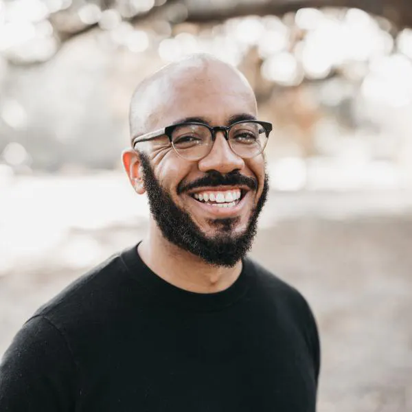 A man with a black ti-shirt and glasses as well as no hair and a beard smiling at the camera