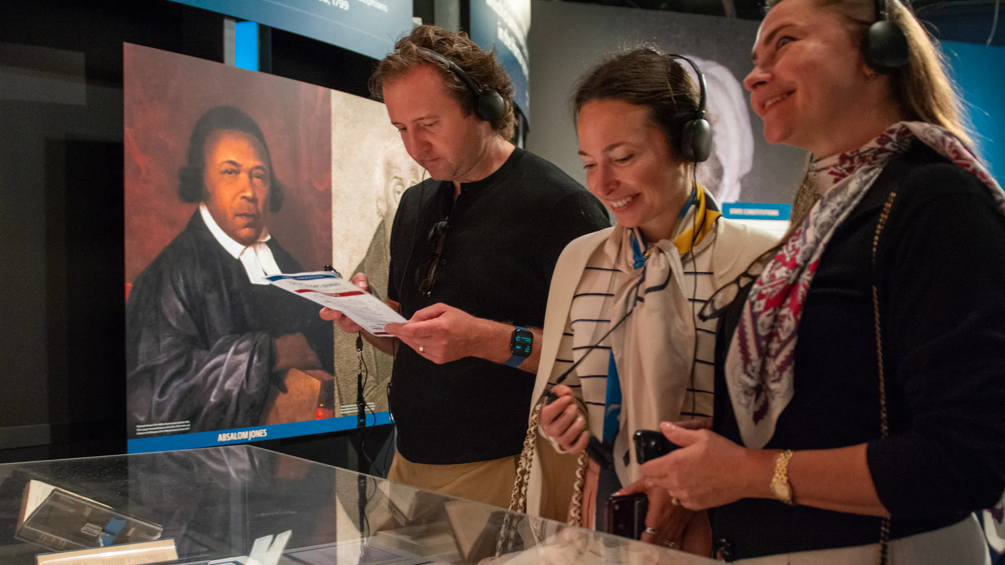 Three people stand in front of a glass case holding documents, wearing headphones