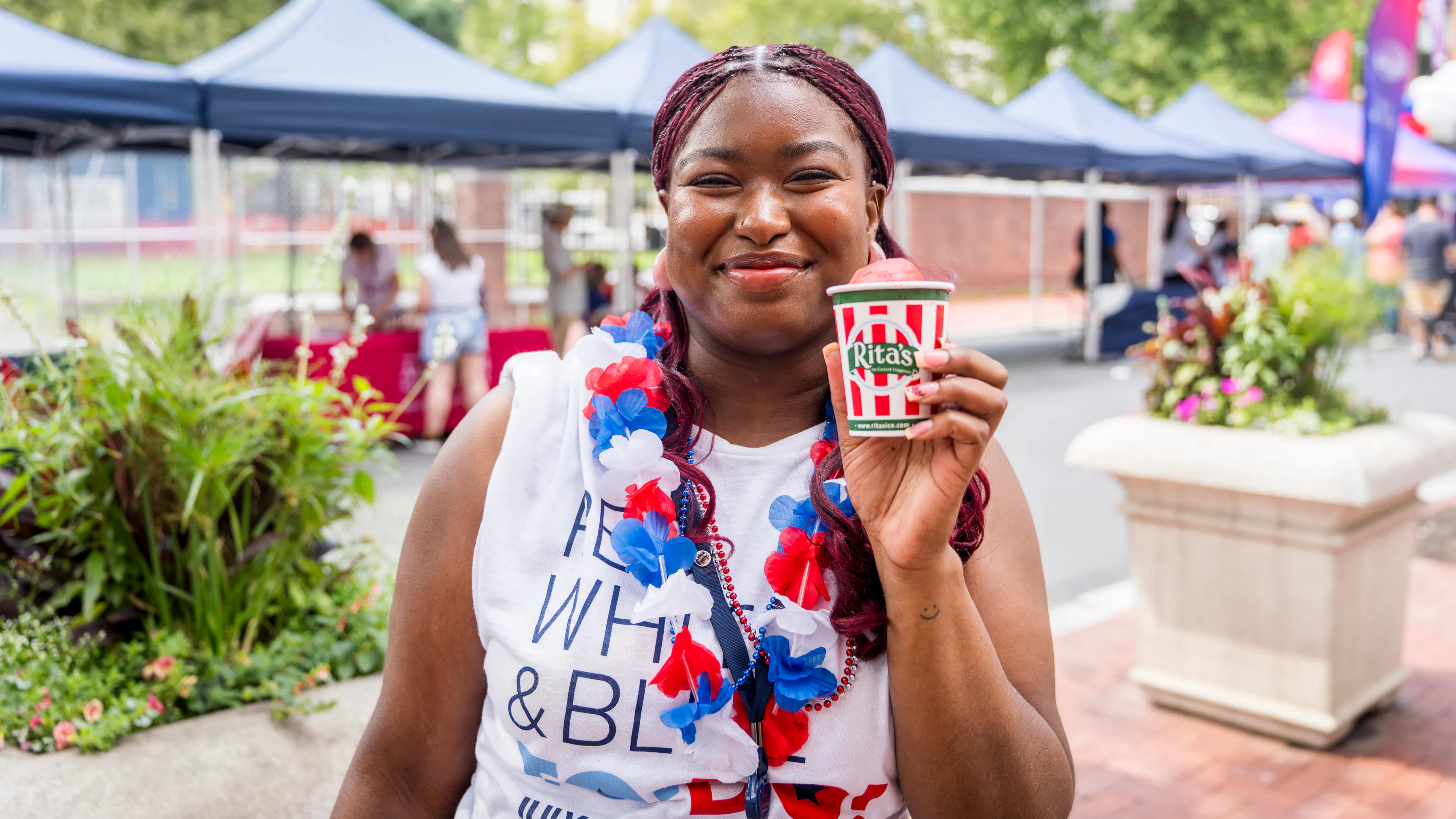 A girl with a white t-shirt and red, white, and blue flowered lei holding a red-and-white striped cup that says Rita's.