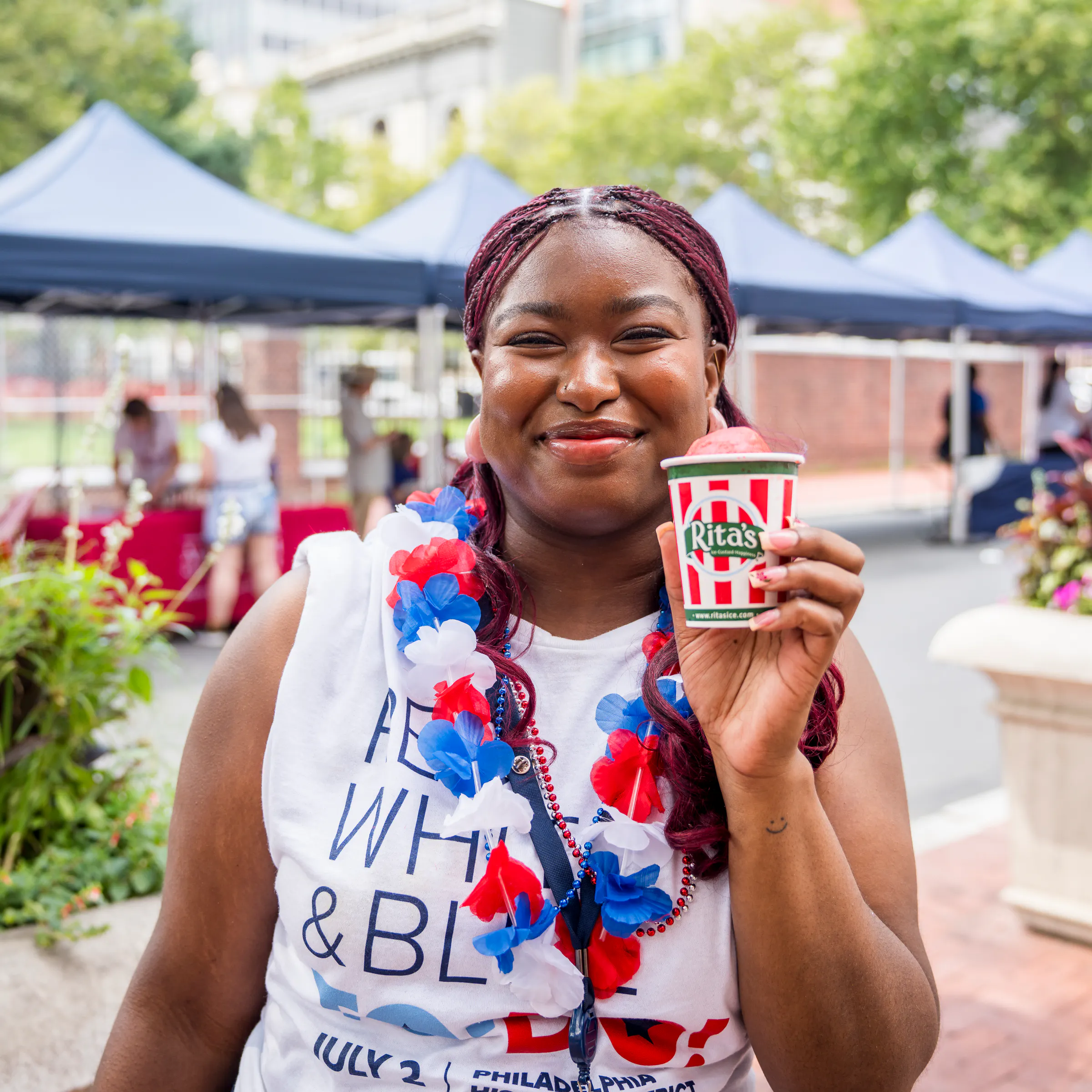 A girl with a white t-shirt and red, white, and blue flowered lei holding a red-and-white striped cup that says Rita's.