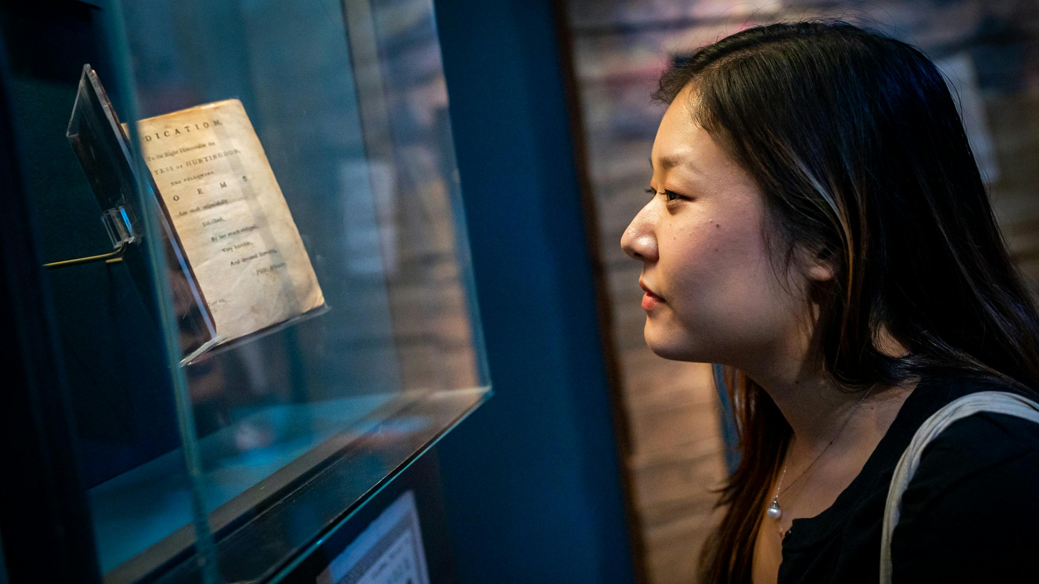 A woman to the right looking closely at a book encased in glass to the left.