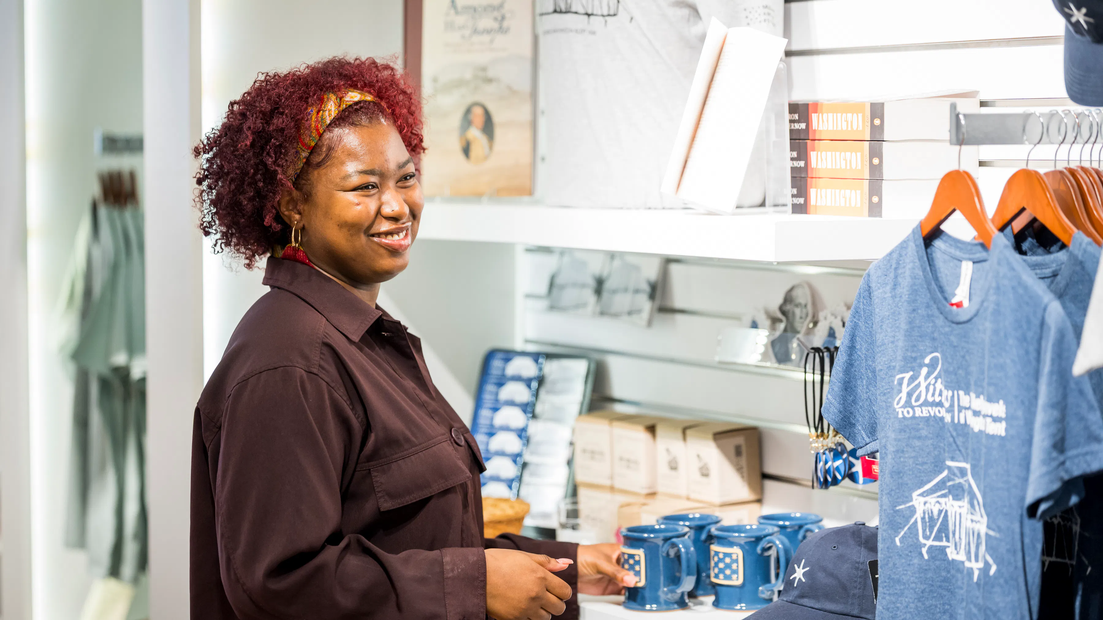 A woman with red curly hair and a brown jacket smiles and looks at blue merchandise including t-shirts, hats and mugs hanging on a white wall.
