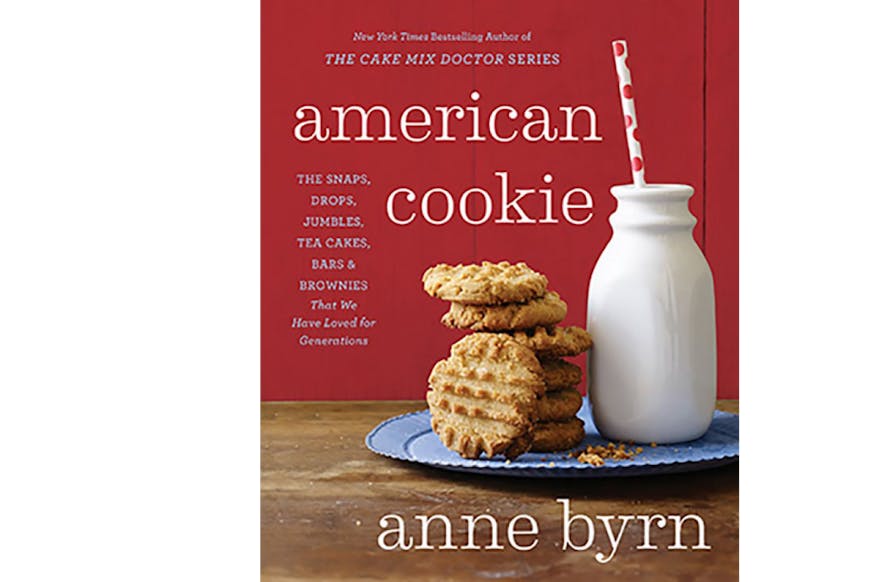 This image depicts the book cover of American Cookie by Anne Byrn. There is a stack of peanut butter cookies and a glass of milk with straw. The milk and cookies are on a blue plate on a wooden table with a red background.