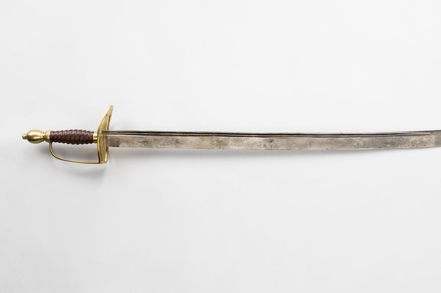 This image is of Benjamin Lincolns sword. The hilt is gold and is on the left side of the image, while the blade is pointing to the right of the image. The sword is resting against a white background.
