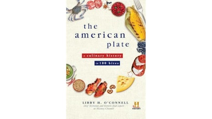 This image shows the book cover of The American Plate: A Culinary History in 100 Bites by Libby O'Connell. The title of the book is written in blue and red. Around the title are items of food forming the shape of a dish. From the top moving to the right is a blue shell grab, meat, anise, a bowl of red berries, a charred corn on the cob in the husk, Swiss cheese, small pretzels, two chicken wings, a bowl of grain, two small red peppers, and a bowl of green soup.