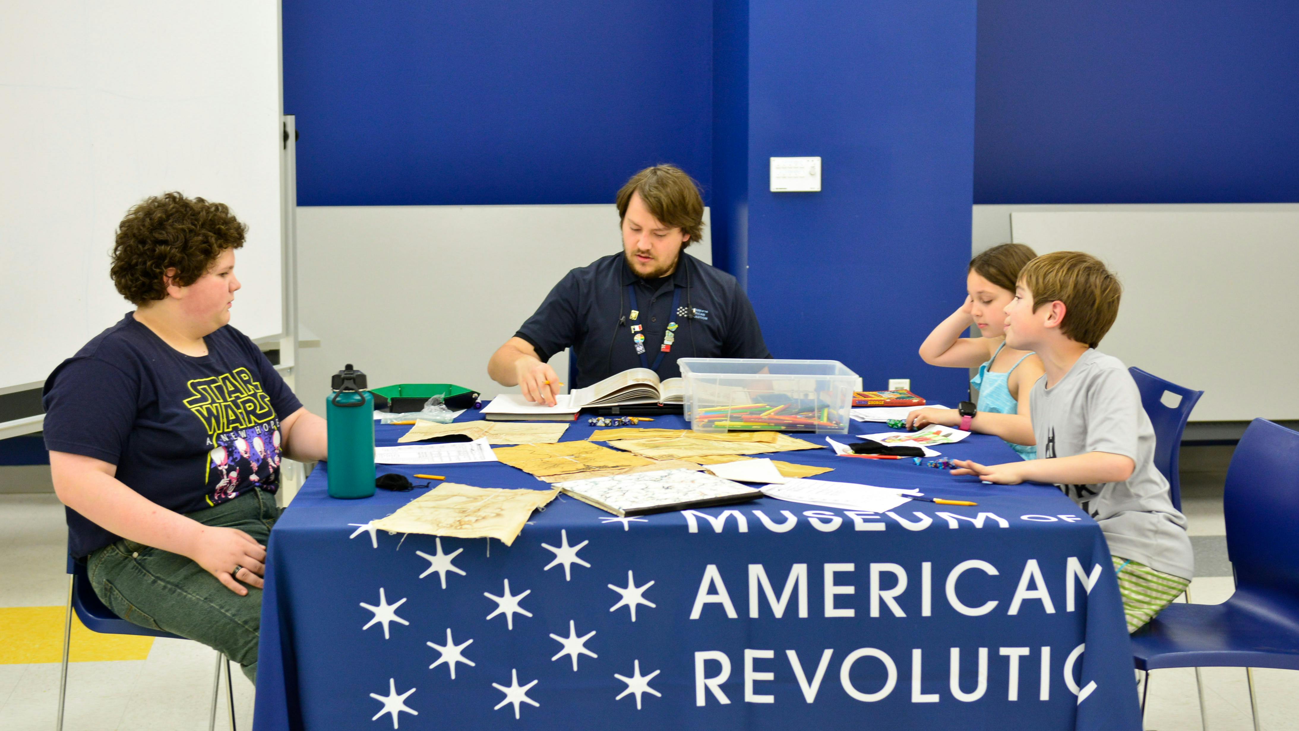 This image shows three young visitors sitting around a table playing a role playing game led by a Museum educator in the center in the Museum's lower level education center.