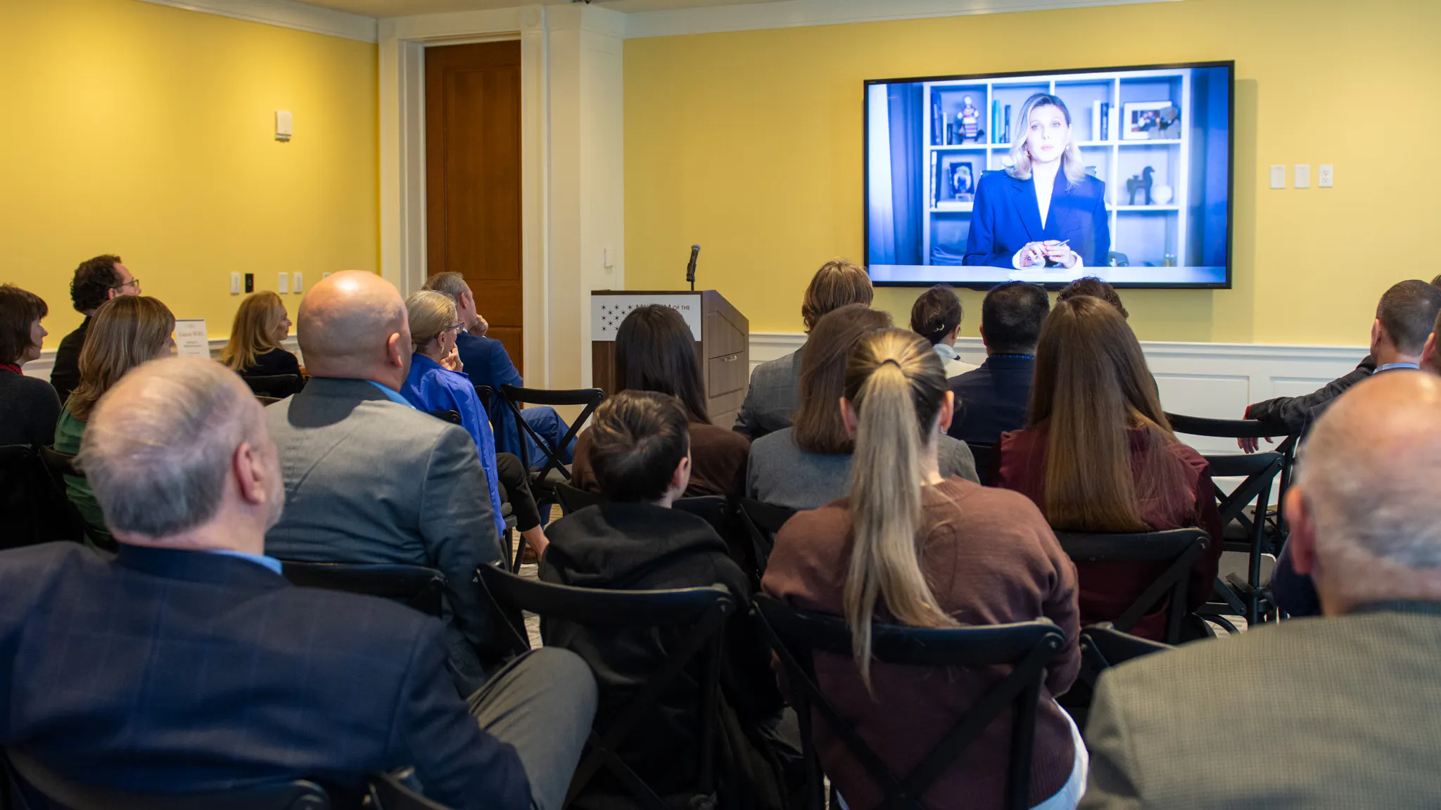 A group of people sit facing a large screen with an image of a seated blond white woman. The room has yellow walls and overhead lighting