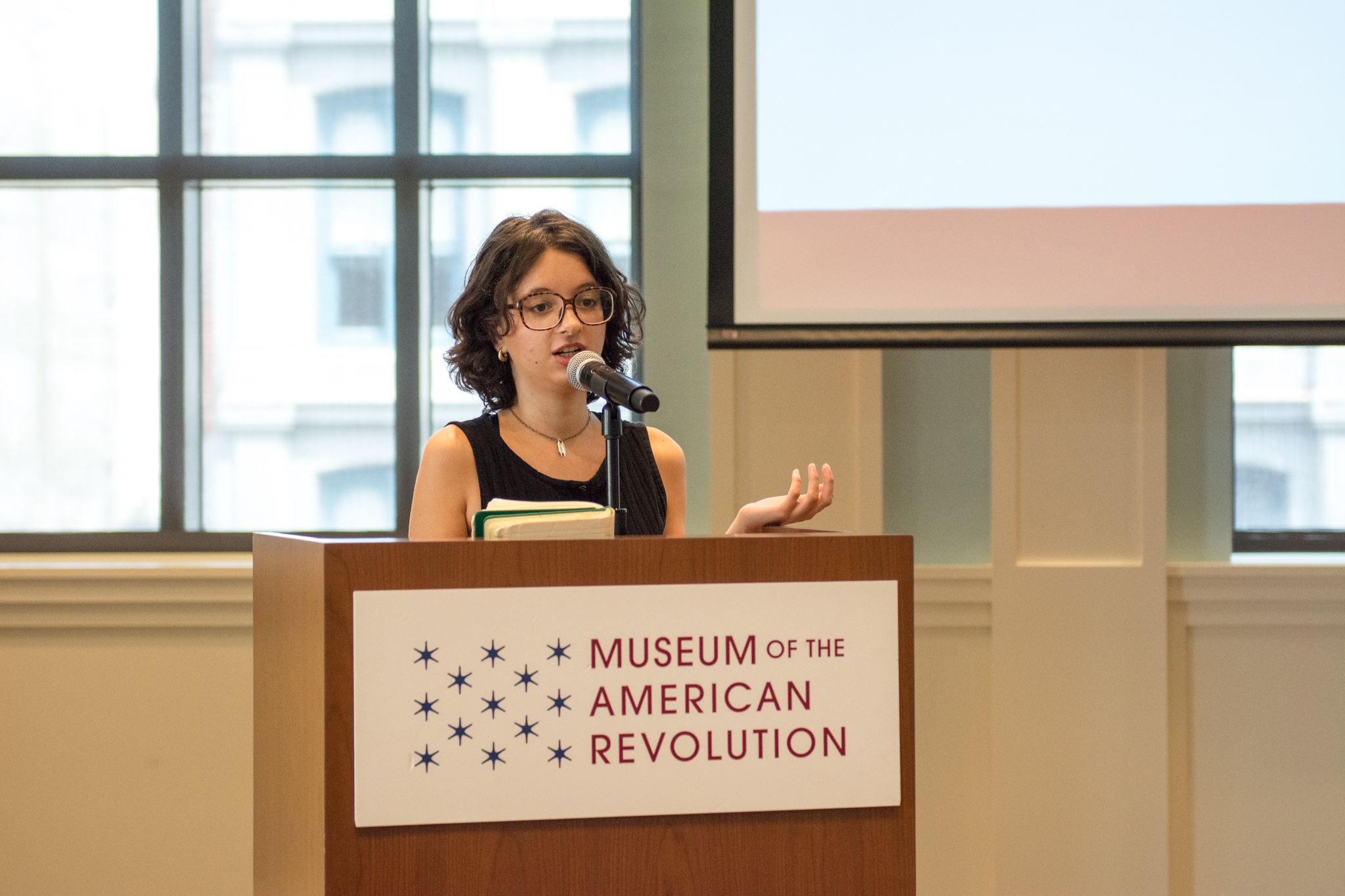 A young white woman with dark hair and glasses speaks at a podium