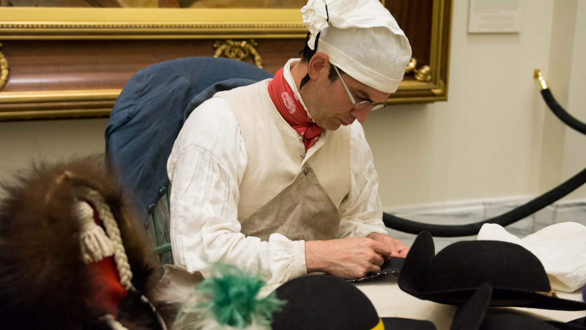 A man in a white 18th-century shirt and cap sewing hats while seated at a table