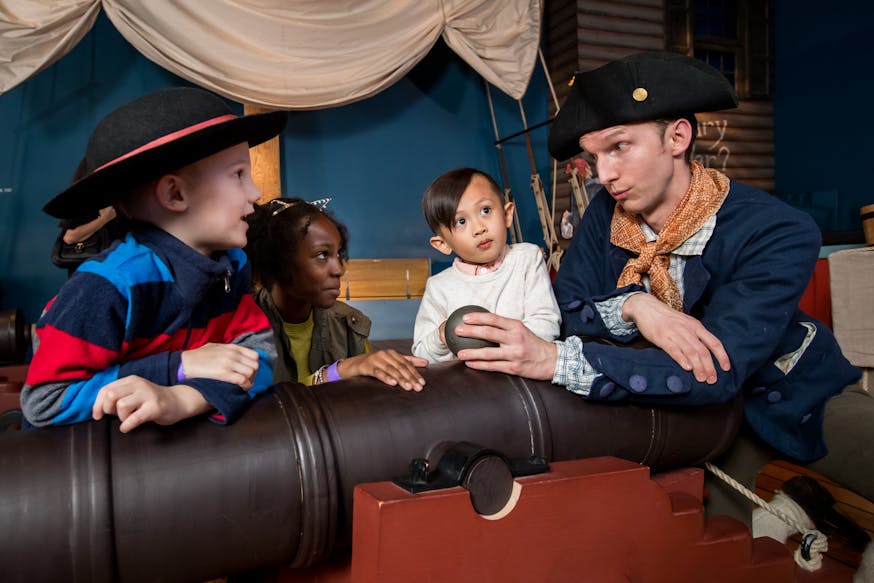 Three children watch a demonstration of a cannon by a costumed Museum educator.