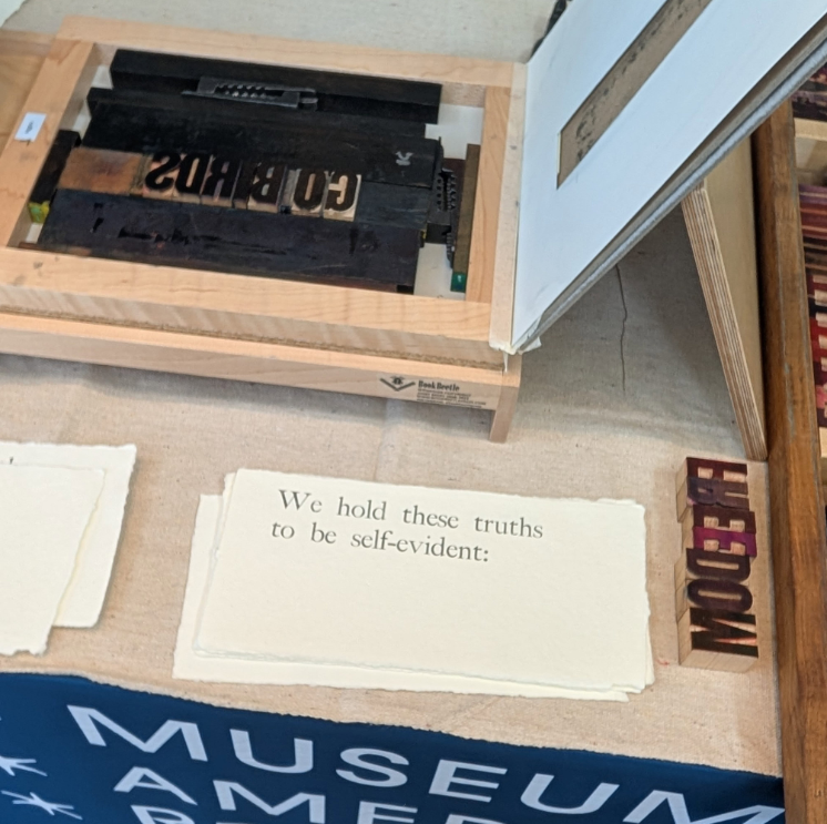 A tabletop printing press next to a typed card that says "We hold these truths to be self-evident."