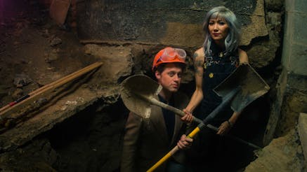 Matt and Melissa Dunphy holding shovels and standing in a hole they dug in the basement of the theater they purchased.