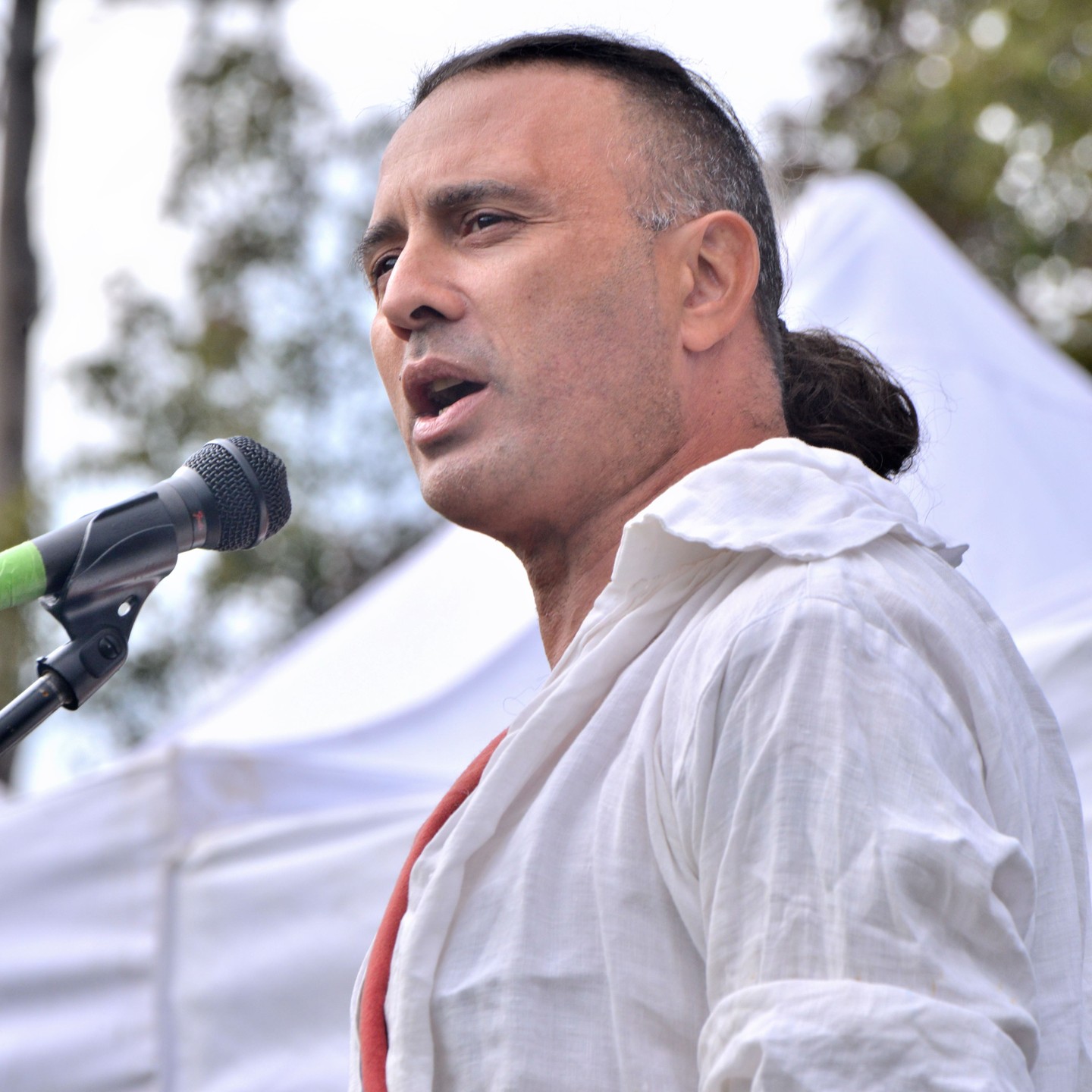 A man with a dark ponytail and white shirt speaks into a microphone