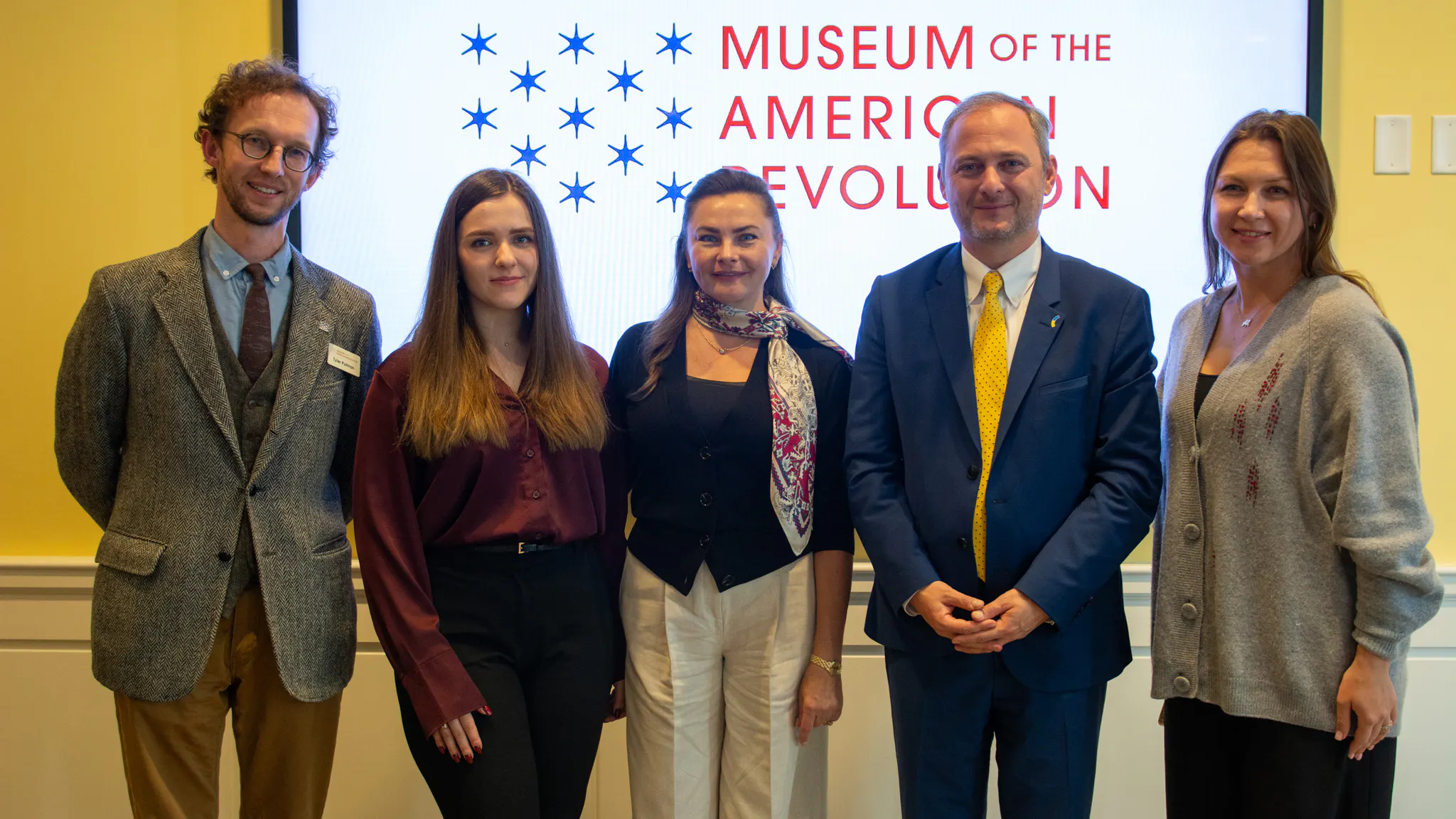 Three women and two men in business attire stand in front of a screen on a yellow wall. The screen displays the Museum's logo