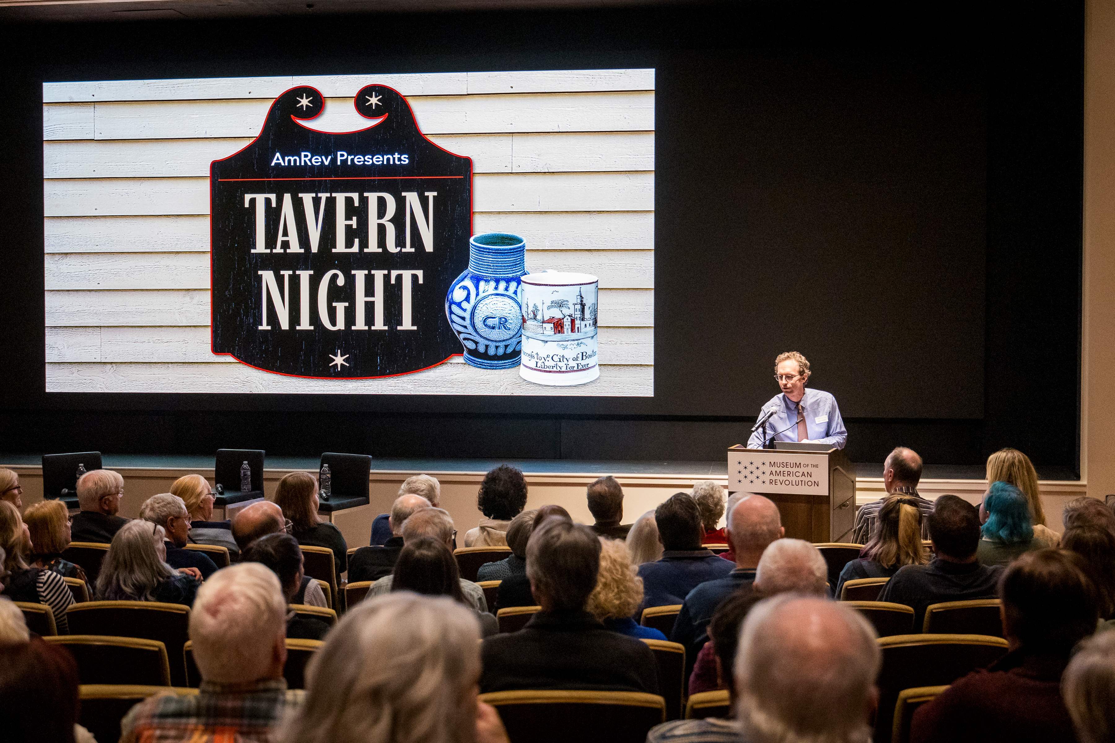 A man speaks at a podium in front of people with a screen behind him that says AmRev Presents Tavern Night