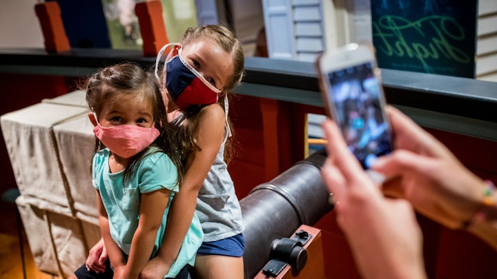 A mother takes a picture of her two young daughters, all wearing masks, on one of the cannons of the Privateer Ship in the galleries.