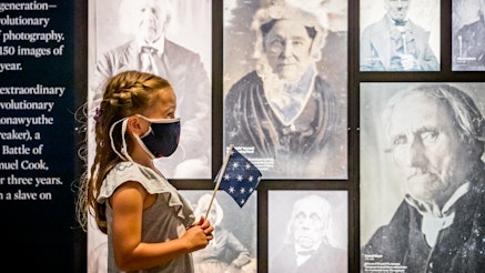 A young, female visitor, wearing a mask and holding a 13-star flag, admires photographs in the Museum's galleries.